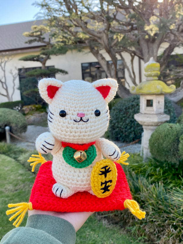 White crocheted maneki, a lucky cat with one of its paws up and a bell around its neck with a gold token near it. It sits atop of a red crocheted cushion, with gold tassels on each corner. It has black beads for eyes and a simplistic smile. Its being held in someones hand outside of a Japanese garden setting.