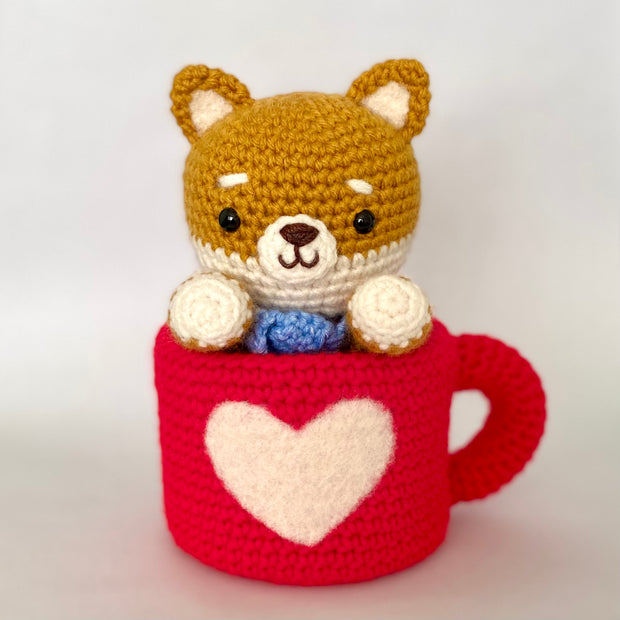 Crocheted plush of a shiba inu dog with a happy face and simple beaded black eyes. It sits inside of a red mug, with a white heart on the front.