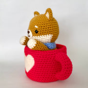 Crocheted plush of a shiba inu dog with a happy face and simple beaded black eyes. It sits inside of a red mug, with a white heart on the front.