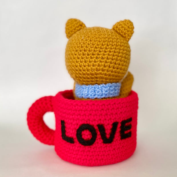 Crocheted plush of a shiba inu dog with a happy face and simple beaded black eyes. It sits inside of a red mug, with "love" written on the back in all black caps.