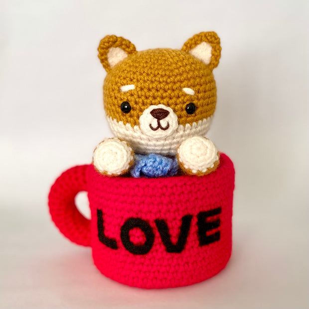 Crocheted plush of a shiba inu dog with a happy face and simple beaded black eyes. It sits inside of a red mug, with "love" written on the back in all black caps.