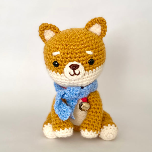 Crocheted plush of a shiba inu dog with a happy face and simple beaded black eyes. It has a blue scarf around its neck and a red collar with a bell on it.
