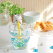 A white ceramic cat with pink ears and gray stripes is draped over a water glass, a yellow straw coming from its mouth. On its back is a brown pot with sprouting mint growing from it.