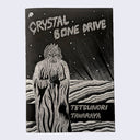 "Crystal Bone Drive" zine cover, silver ink illustration of a alien bearded character in a flowing robe on textured floor with light beams coming from behind. 