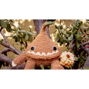 A silly looking brown plush doll with a teardrop shaped body and a green sprout growing from its head. It ha a large underbite smile that takes up most of its face and has arms and legs coming out of the head. It holds a faded yellow rose in its hand.