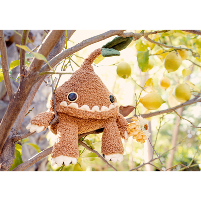 A silly looking brown plush doll with a teardrop shaped body and a green sprout growing from its head. It ha a large underbite smile that takes up most of its face and has arms and legs coming out of the head. It sits on a lemon tree.