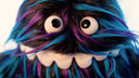 Close up of plush doll of a monster creature, similar to a muppet, with a long underbite smile that wraps around its face and long black fur with blue and purple color highlights. 