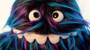 Close up of plush doll of a monster creature, similar to a muppet, with a long underbite smile that wraps around its face and long black fur with blue and purple color highlights. 