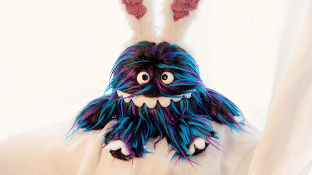 Close up of Plush doll of a monster creature, similar to a muppet, with a long underbite smile that wraps around its face and long black fur with blue and purple color highlights. It has wears a headband with white fluffy bunny ears on its head.