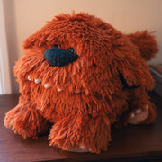 Rounded long haired plush dog, with an exaggerated large nose and many rounded teeth. Its fur is a reddish brown.