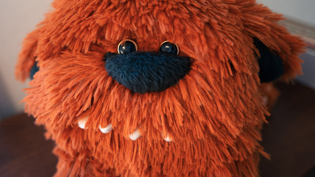 Rounded long haired plush dog, with an exaggerated large nose and many rounded teeth. Its fur is a reddish brown.