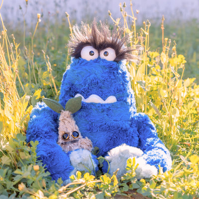 2 fluffy plush creatures sitting outside. The larger plush is blue with an underbite, large eyes and very hairy eyebrows. Resting in his lap is a small brown plush with a sprout top and circular eyes.