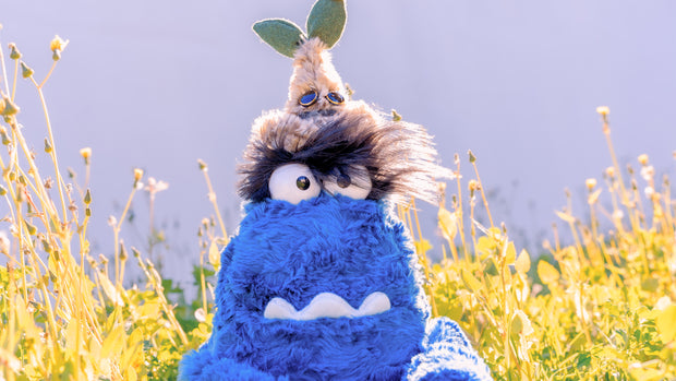 2 fluffy plush creatures sitting outside. The larger plush is blue with an underbite, large eyes and very hairy eyebrows. Resting atop his head is a small brown plush with a sprout top and circular eyes.