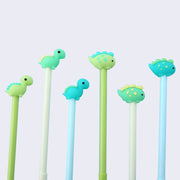 Close up of 6 blue, green and white color way pens with rubber chibi dinosaur pen toppers. Dinosaurs include Brachiosaurus (blue spikes on light green, light green spikes on green, and green spikes on teal blue) and Stegosaurus (blue spikes/dots on light green, yellow spikes/dots on dark green, and green spikes/dots on teal blue) 