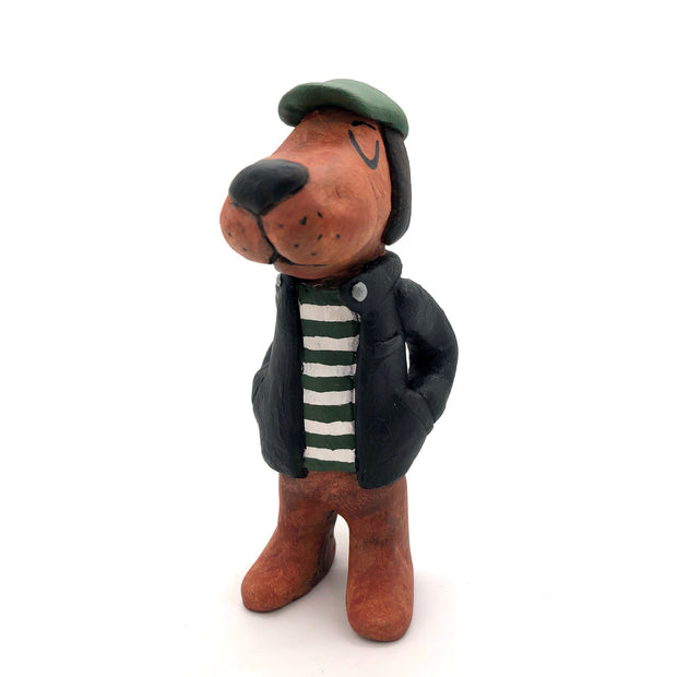Painted clay sculpture of a brown dog, standing up on 2 legs like a human with its hands in its jacket pocket. It wears a black jacket with a striped shirt and a green newsboy cap. 