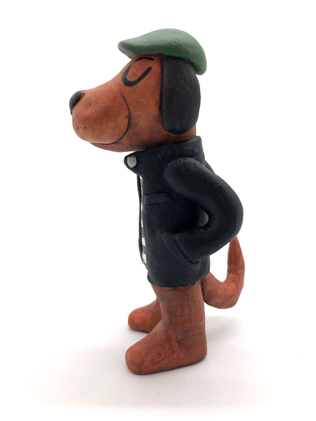 Painted clay sculpture of a brown dog, standing up on 2 legs like a human with its hands in its jacket pocket. It wears a black jacket with a striped shirt and a green newsboy cap.