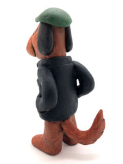 Painted clay sculpture of a brown dog, standing up on 2 legs like a human with its hands in its jacket pocket. It wears a black jacket with a striped shirt and a green newsboy cap.