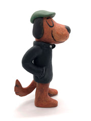 Painted clay sculpture of a brown dog, standing up on 2 legs like a human with its hands in its jacket pocket. It wears a black jacket with a striped shirt and a green newsboy cap.