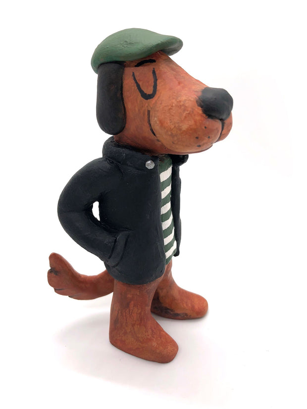 Painted clay sculpture of a brown dog, standing up on 2 legs like a human with its hands in its jacket pocket. It wears a black jacket with a striped shirt and a green newsboy cap.