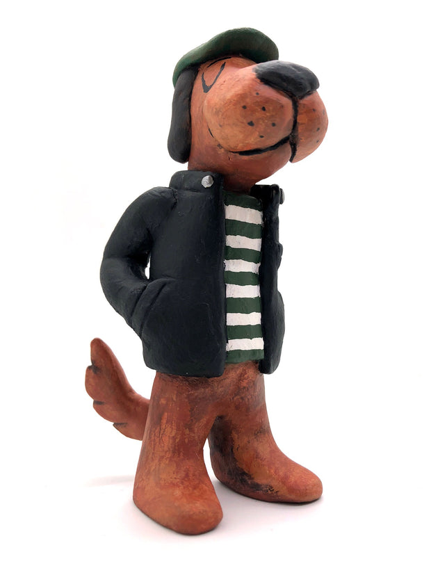 Painted clay sculpture of a brown dog, standing up on 2 legs like a human with its hands in its jacket pocket. It wears a black jacket with a striped shirt and a green newsboy cap.