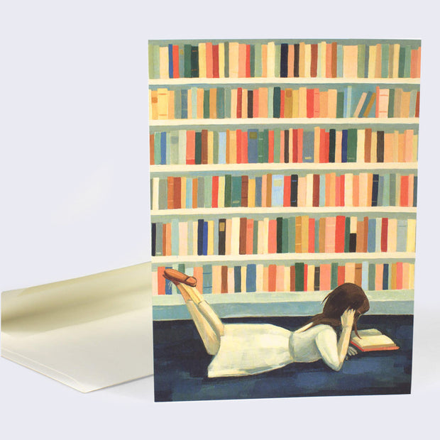 The front of note card is shown with a white envelope behind it. The notecard has an illustration of a girl laying on the floor reading a book in the library.
