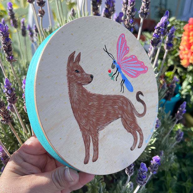 Round wooden panel with a painted dog, standing and looking over its shoulder at a large pink butterfly which holds out a flower for the dog. Edges of the panel are painted teal.