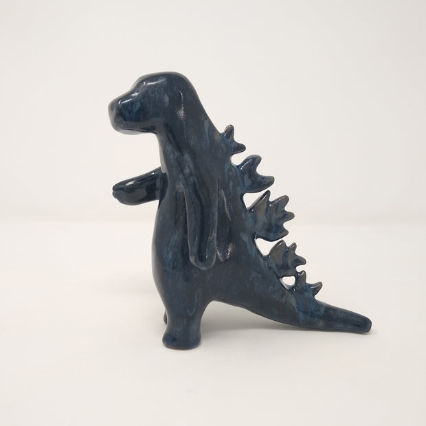 Ceramic sculpture of a dark blue abstract Godzilla figure, with abstract spikes on its back, no facial features, and one arm extended out.