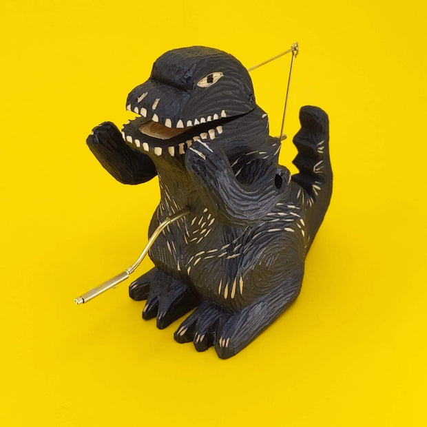 Carved wooden sculpture of a black Godzilla, standing with arms propped up, mouth slightly ajar, and crank on its stomach.