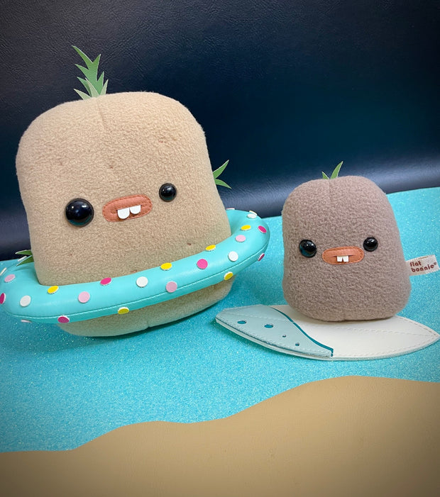 Set of 2 plush potatoes, one light brown and the other smaller and medium brown. They have mismatched eyes, buckteeth and sprouts growing from their heads. Larger one has a pool ring floaty and the baby has a surfboard.