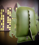 Back view of a green vinyl plush, shaped as a Big Boss Robot figure with spikes on its back and a tail. A paper cut out city is in the background.