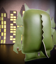 Back view of a green vinyl plush, shaped as a Big Boss Robot figure with spikes on its back and a tail. A paper cut out city is in the background.