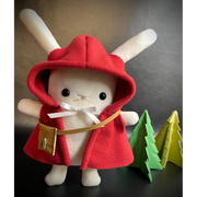 Plush of a white bunny with long, skinny ears and outward facing arms. It wears a red hooded cloak with a white bow around the neck and a small leather crossbody pouch. Behind are 2 green pine trees made out of fabric.