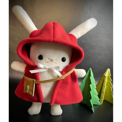 Plush of a white bunny with long, skinny ears and outward facing arms. It wears a red hooded cloak with a white bow around the neck and a small leather crossbody pouch. Behind are 2 green pine trees made out of fabric.