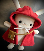 Plush of a white bunny with long, skinny ears and outward facing arms. It wears a red hooded cloak with a white bow around the neck and a small leather crossbody pouch.