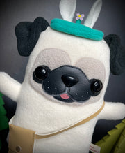 Plush sculpture of a pug, standing on 2 legs with its arms extended out. It wears a small crossbody satchel and a teal blue headband that has a small flower springing up from it.
