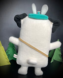 Plush sculpture of a pug, standing on 2 legs with its arms extended out. It wears a small crossbody satchel and a teal blue headband that has a small flower springing up from it.
