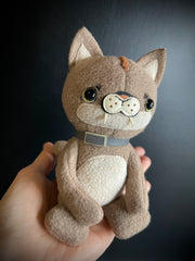 Plush sculpture of a tan puma cub, sitting in the palm of someone's hand with a gray color around its neck.