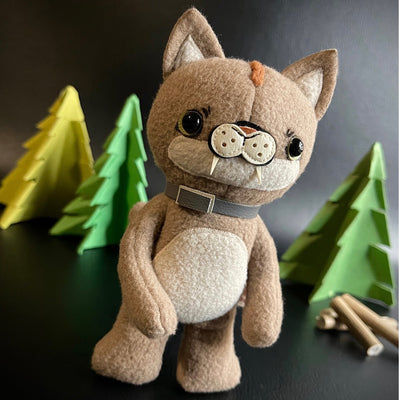 Plush sculpture of a tan puma cub, standing on just 2 legs with a gray collar around its neck and black bead eyes. In the background are foam pine trees and logs.
