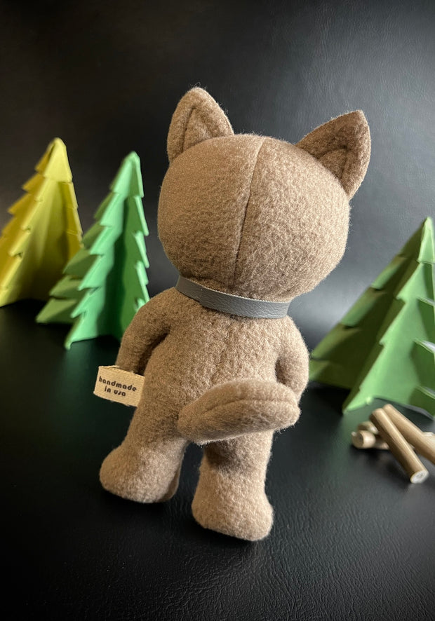 Back view of plush sculpture of a tan puma cub, standing on just 2 legs with a gray collar around its neck and a mid length tail. In the background are foam pine trees and logs.