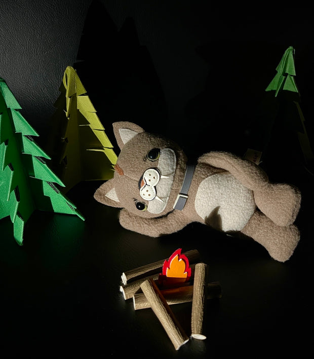 Plush sculpture of a tan puma cub, laying on its side with its arm supporting its head. It wears a gray collar around its neck and lays in front of a campfire with felt pine trees in the background.