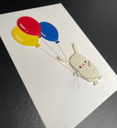 Vinyl sewn art piece, with slightly raised 3D elements of a small white rabbit holding a bundle of 3 balloons, blue, yellow and red. Shown at an angle to display 3 dimensionality.