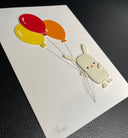 Vinyl sewn art piece, with slightly raised 3D elements of a small white rabbit holding a bundle of 3 balloons, orange, yellow and red. Shown at an angle to show three dimensionality.