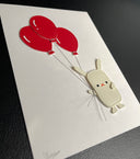 Vinyl sewn art piece, with slightly raised 3D elements of a small white rabbit holding a bundle of 3 red balloons. Shown at an angle to see the three dimensionality.