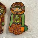 Illustrated wood cut human in a wearing a full head mask of a monster, with a small peek of the person showing through. He sits on his knees and wears a wildly patterned orange and green outfit.