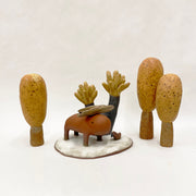 Scene of several earth tone ceramic pieces. A burnt orange colored capybara like figure stands with a small branch on its head. Behind is an abstract spiky black and brown tree, a small bird sits nearby on the shared platform. Off the platform are 3 abstract round trees.  