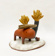 Scene of several earth tone ceramic pieces. A burnt orange colored capybara like figure stands with a small branch on its head. Behind is an abstract spiky black and brown tree, a small bird sits nearby on the shared platform.