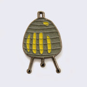 Enamel pin of a mid century gray paper table lamp on 3 legs, with yellow accents acting as light.