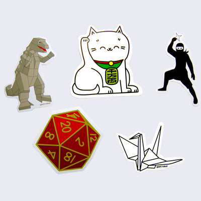 5 different designs of white bordered die cut stickers. Stickers include: geometric Godzilla made out of cardboard, a classic Japanese maneki cat, a ninja dressed in all black holding a throwing star, a red 20 sided die, and an origami crane.