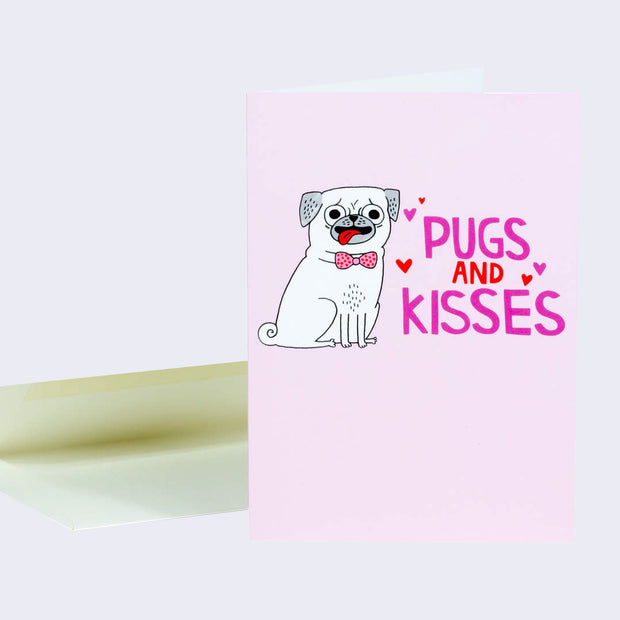 Pink greeting card with an illustration of a happy goofy pug with its tongue out. "Pugs and Kisses" is written in pink and red font with little hearts floating nearby.