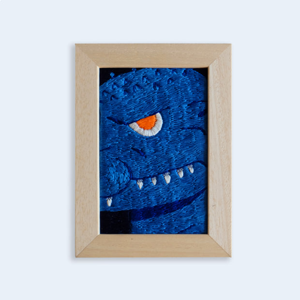 Embroidery piece in a thick light stained wooden frame. Embroidery is a close up shot of a deep blue Godzilla, with a single orange eye looking at the viewer menacingly and white teeth showing out of a closed mouth. 